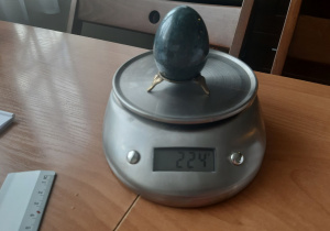 na wadze kuchennej stoi ciało stałe o kształcie nieregularnym na podstawce, waga wskazuje masę jedynie ciała o kształcie nieregularnym, ciałem tym jest ozdobne jajko wykonane z ceramiki